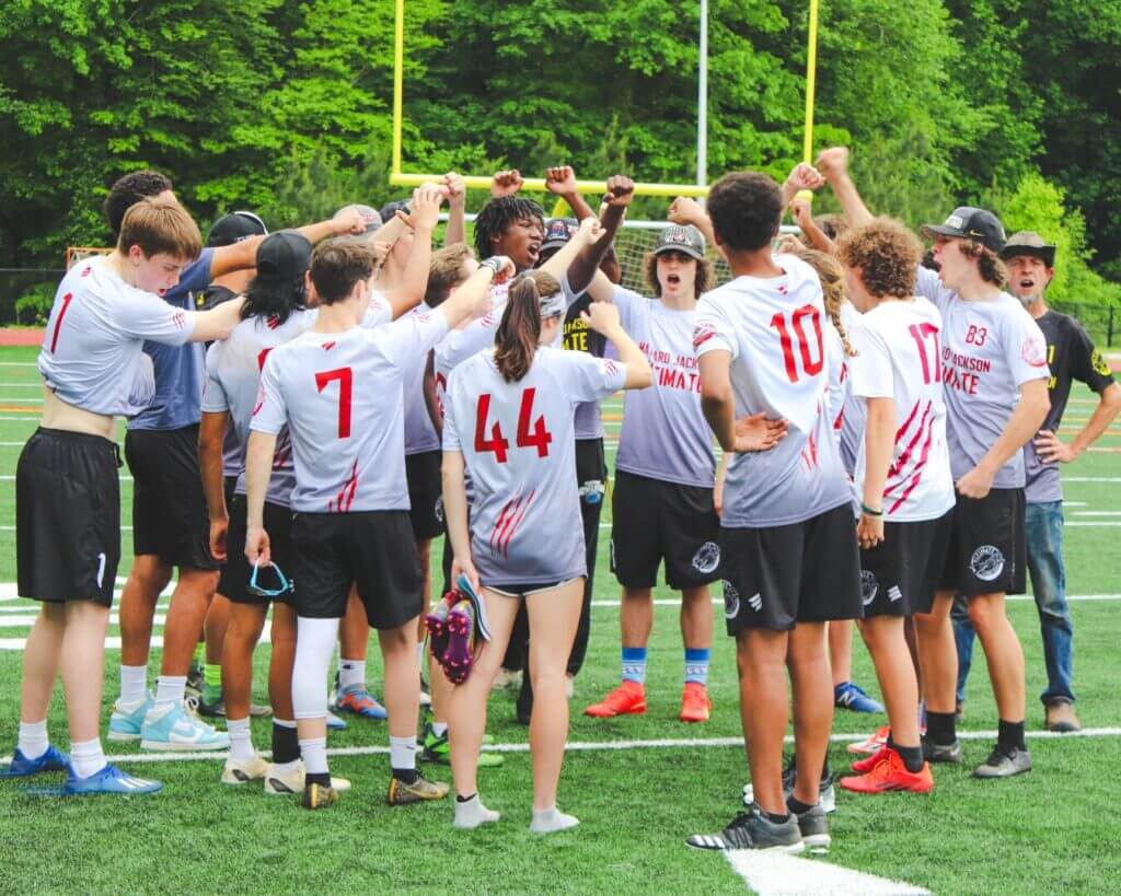A group of ultimate players in matching gray jerseys huddle on a turf field for a game.